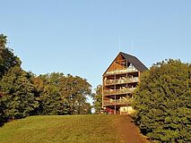 Die VHS befindet sich in der Kolpingstraße hinter dem Flederhaus St. Pölten. Blick über ein herbstliches Feld auf einen Berg mit Wald und einer Lichtung: Darauf ein mehrstöckiges Holzhaus - das Flederhaus St. Pölten.