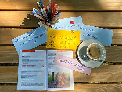 Ein Stilleben von oben fotografiert: Eine Kaffeetasse mit Kaffee gefüllt, 5 blaue Notizzettel, ein aufgeschlagenes Programmheft, ein Glas mit verschiedenen Farbstiften. Mittig eine gelbe Karte mit der handschriftlichen Notiz "Was wünscht sich St. Pölten von ihrer VHS?"